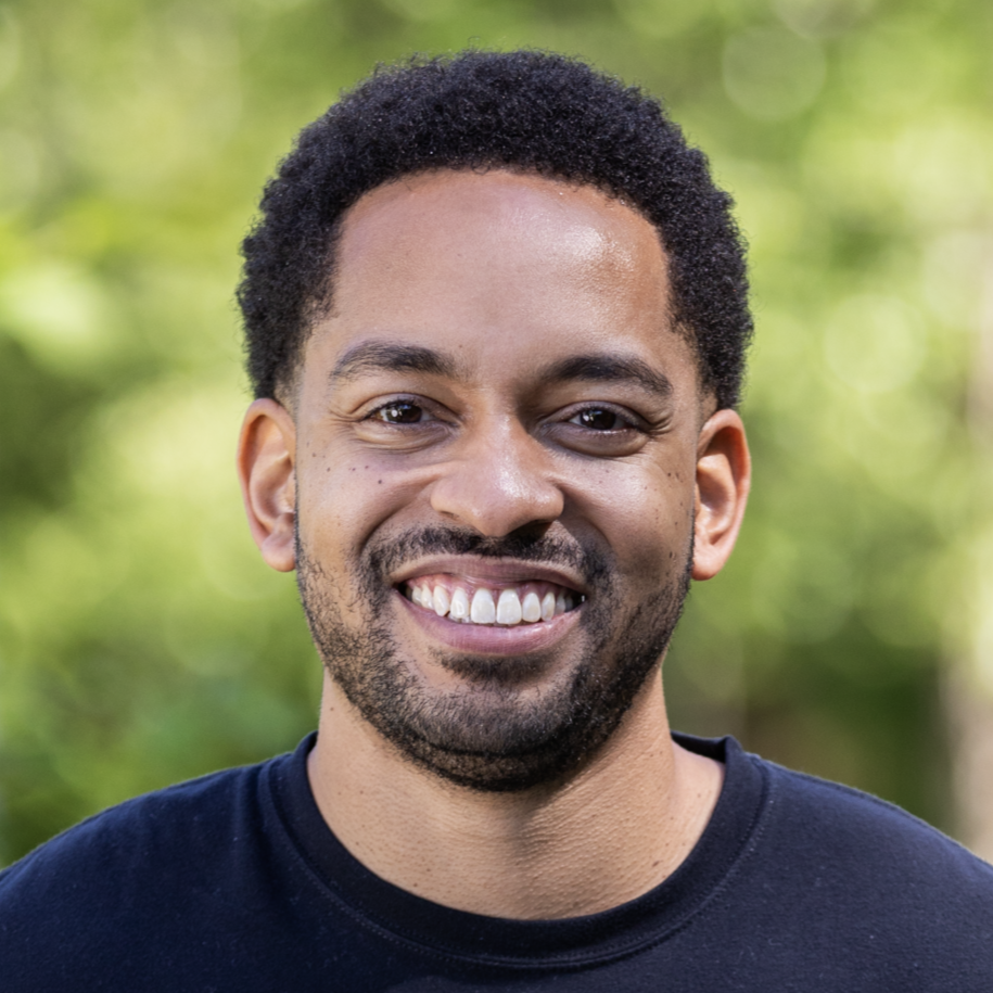 Martez smiling and wearing a black shirt in front of a lush green background
